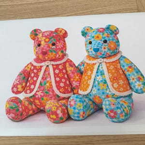 Könnte beinhalten: Zwei handgefertigte Teddybären mit Blumenmustern. Einer ist rot mit weißem Kragen, der andere blau mit orangefarbenem Kragen. Beide haben Knopfverschlüsse und sitzen auf einer weißen Oberfläche.