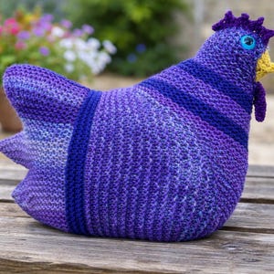 Könnte beinhalten: Ein handgefertigtes, gestricktes Huhn in Lila- und Blautönen. Das Huhn hat einen detaillierten Kamm, einen Kehllappen und ein blaues Auge. Der Körper weist Streifenmuster auf und das Stück ist auf einer Holzoberfläche platziert, was auf einen Dekorationsartikel hindeutet.