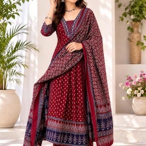 May include: A deep maroon Anarkali suit featuring a white geometric print, a V-neckline, and three-quarter sleeves. The ensemble includes matching wide-leg trousers and a long, coordinating dupatta with a dark blue border and intricate floral patterns.