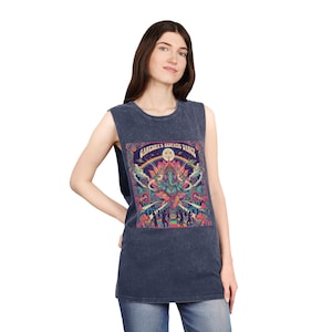 May include: A sleeveless, dark blue top featuring a colorful graphic of a deity with the text "GANESHA'S ECSTATIC DANCE". The graphic includes a rainbow, sun, and figures. The top is worn with blue jeans.