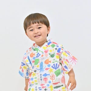May include: A child wearing a white kimono-style robe with a colorful sea life pattern. The robe features illustrations of fish, starfish, and other marine creatures in various colors. The robe is tied at the waist.
