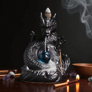 Op de afbeelding: Een zwarte keramische draken wierookbrander met een blauwe bol en een kegelvormige wierook. Rook stroomt uit de bek van de draak, wat een mystiek effect creëert. Wierookstokjes en een kleine kom zijn ook zichtbaar.