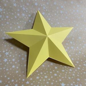 May include: A large, five-pointed yellow paper star. The star is folded with sharp angles and sits on a tan background with a pattern of white stars. The star's shadow is visible.