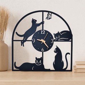 Puede incluir: Reloj de pared de metal negro con diseño de gatos. El reloj tiene una esfera azul oscuro con manecillas doradas y está rodeado de siluetas de gatos negros. Un gato intenta atrapar una mariposa. Aproximadamente 30 cm de diámetro.