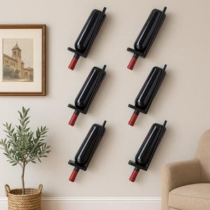 Puede incluir: Seis botellas de vino negro se exhiben en soportes de pared. Las botellas tienen vino tinto visible y tapones rojos. Los soportes son negros y están montados en una pared de color neutro. Una imagen enmarcada y una pequeña planta están en el fondo.