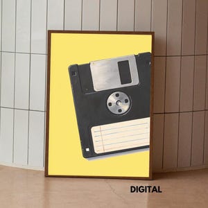 Könnte beinhalten: Ein gerahmter rechteckiger Druck mit einer schwarzen Diskette vor einem einfarbigen gelben Hintergrund. Die Diskette hat einen silbernen Metallschieber, eine kreisförmige Nabe in der Mitte und ein weißes rechteckiges Etikett mit blauen und roten horizontalen Linien. Das Wort DIGITAL ist unten rechts in Schwarz aufgedruckt.