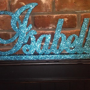 May include: A teal glitter name sign with the name "Isabella" in cursive script. The sign is made of wood and has a rustic look.