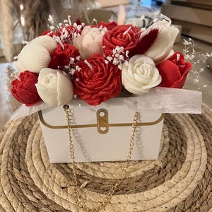 Può includere: Una composizione floreale decorativa con candele a forma di rose rosse e bianche, tulipani e candele a forma di conchiglia. Le candele sono in una scatola bianca con un manico a catena dorata, su un tappetino intrecciato.