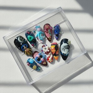 May include: A collection of ten intricately designed artificial nails, each with a unique theme. Designs include a cartoon character, an octopus, and a night sky scene. The nails are displayed in a clear plastic case, showcasing the detailed artistry.