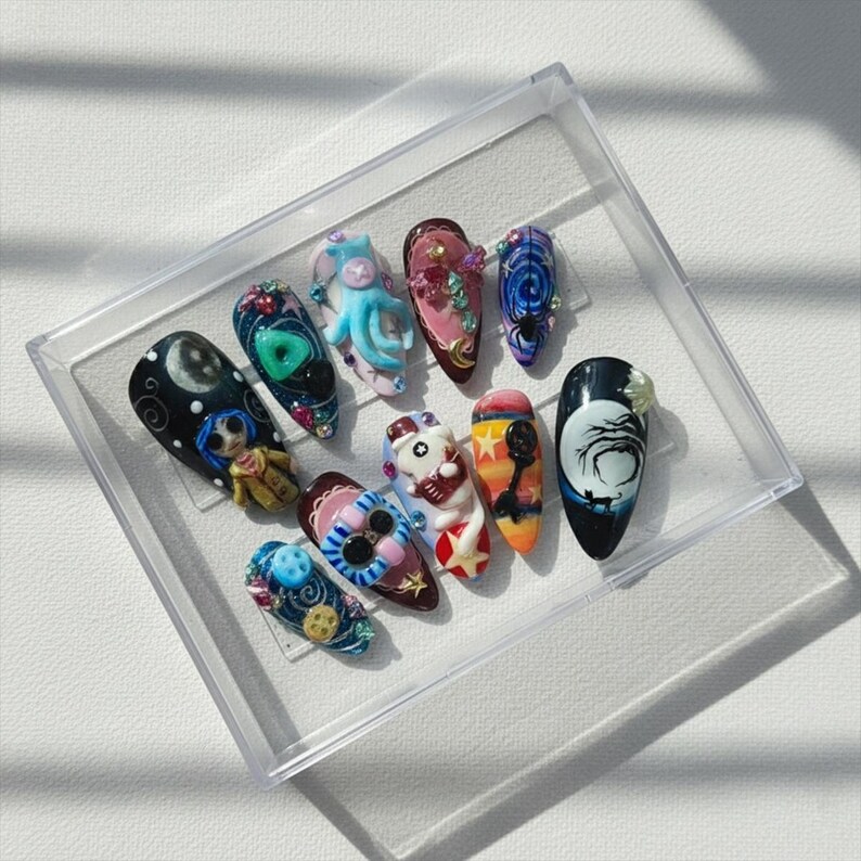 May include: A collection of ten intricately designed artificial nails, each with a unique theme. Designs include a cartoon character, an octopus, and a night sky scene. The nails are displayed in a clear plastic case, showcasing the detailed artistry.
