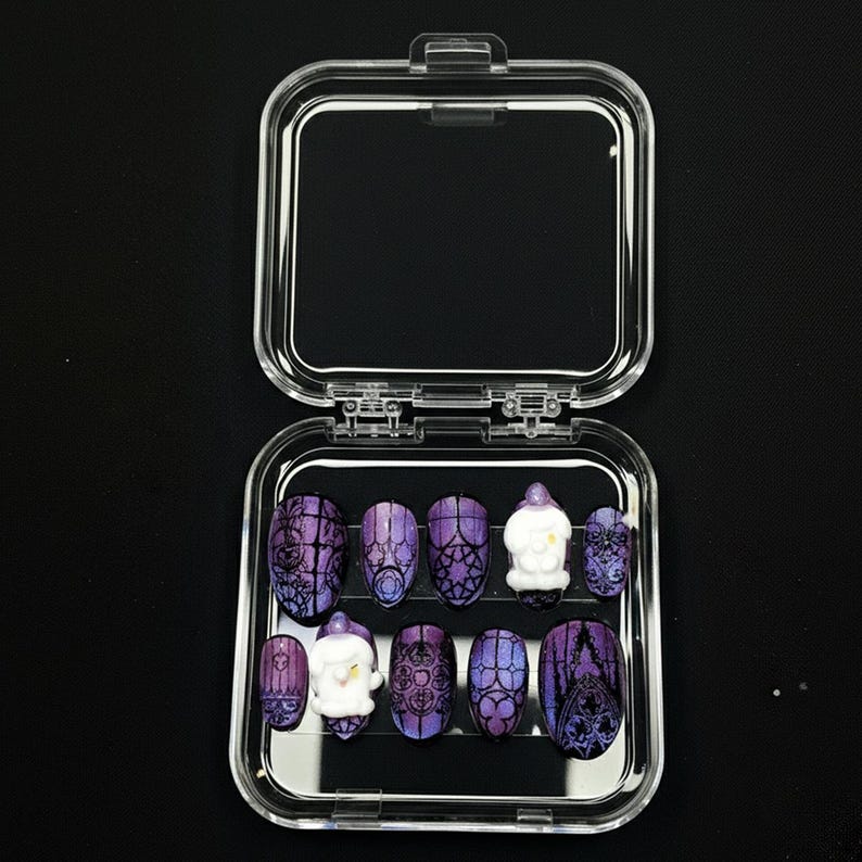 May include: A set of press-on nails in a clear plastic case. The nails feature a purple and black design with a gothic aesthetic. Some nails have a white, snowman-like embellishment. The nails are oval-shaped.