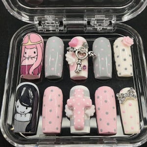 Könnte beinhalten: Ein Set aus zehn künstlichen Nägeln in einer transparenten Kunststoffbox. Die Nägel weisen verschiedene Designs auf, darunter Zeichentrickfiguren, Polka Dots und dekorative Verzierungen wie ein Schlüssel, eine Schleife und ein Kreuz. Farben sind Pink, Grau, Weiß und Schwarz.