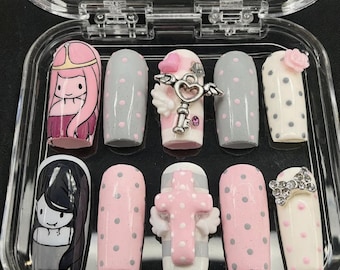 May include: A collection of ten false nails presented in a clear case. The nails showcase diverse designs, such as cartoon characters, polka dots, and decorative elements like a key, a bow, and a cross. The colour palette includes pink, grey, white, and black.