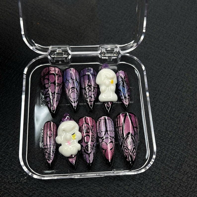 May include: A set of ten long, pointed false nails in a clear plastic case. The nails feature a gradient of purple and black with a gothic design. Two nails have a white ghost-like charm with yellow accents.