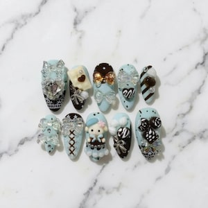 May include: Ten artificial nails, each painted a light blue colour. The nails are decorated with bows, hearts, pearls, and miniature figurines. Some nails have lace patterns and sparkling crystals. The nails are arranged on a marble surface.