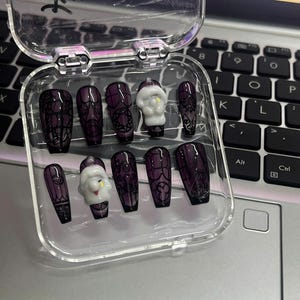 May include: A set of dark purple press-on nails with black lace and gothic designs, including a small white Santa Claus nail. The nails are displayed in a clear plastic case, perfect for a spooky or holiday look.