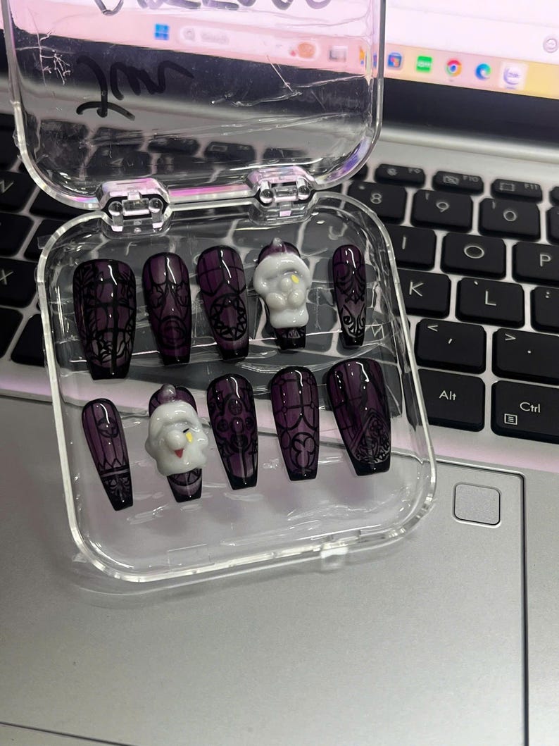 May include: A set of dark purple press-on nails with black lace and gothic designs, including a small white Santa Claus nail. The nails are displayed in a clear plastic case, perfect for a spooky or holiday look.