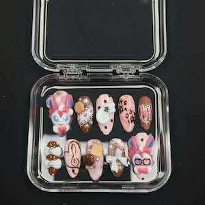 Może przedstawiać: Zestaw dziesięciu sztucznych paznokci w przezroczystym plastikowym etui. Paznokcie zdobią różne wzory, w tym postacie z kreskówek, nuty i elementy dekoracyjne w odcieniach różu, brązu i bieli. Każdy paznokieć ma unikalny wzór.