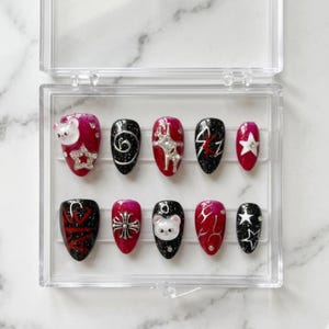 May include: A set of ten press-on nails in a clear plastic case. The nails feature various designs, including a white bear, stars, swirls, and a cross, with colours of red, black, and silver glitter.