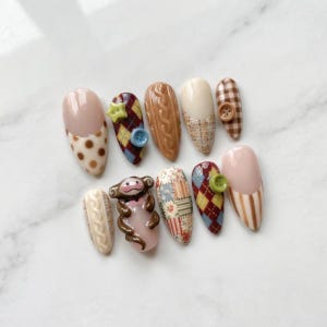 May include: A set of ten almond-shaped false nails with various designs. The designs include polka dots, argyle patterns, cable knit, tweed, patchwork, and a monkey figurine. Colors include beige, brown, red, blue, and green. Some nails feature buttons.