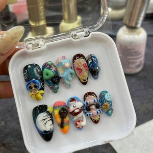 May include: A collection of ten intricately designed artificial nails, each with a unique, colorful, and detailed design. The nails feature various themes, including cartoon characters, abstract patterns, and 3D embellishments, presented in a white tray.