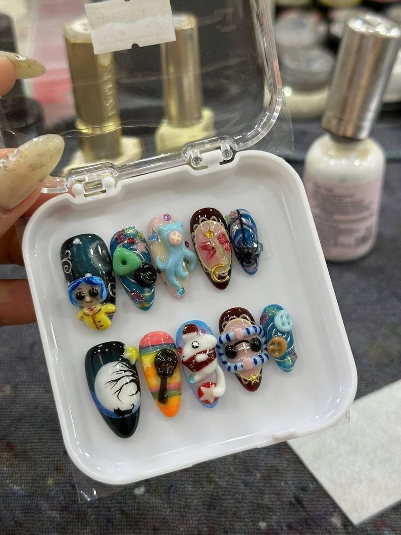 May include: A collection of ten intricately designed artificial nails, each with a unique, colorful, and detailed design. The nails feature various themes, including cartoon characters, abstract patterns, and 3D embellishments, presented in a white tray.
