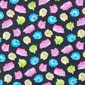 May include: Black fabric with a repeating pattern of cartoon pigs in various colors, including pink, blue, yellow, and green. The pigs have floral accents and are scattered across the surface.