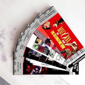 Pode incluir: Um conjunto de bilhetes para o concerto "The Celebration Tour" de Madonna. Os bilhetes estão em leque, com fundo vermelho e imagens de Madonna. Informações sobre o local e a data também são visíveis.