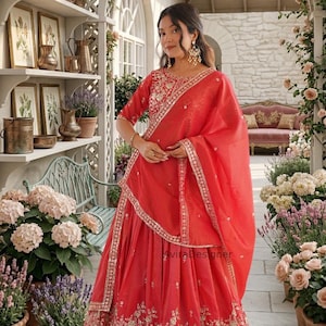 May include: A striking red Indian outfit, comprising an embroidered top and skirt, and a matching scarf. The outfit is decorated with gold-coloured detailing. The image is set in a garden with floral arrangements and a stone wall.