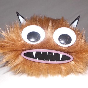 May include: A whimsical, brown furry monster with large googly eyes and a wide, toothy grin. The creature has small black and white ears, creating a playful and cartoonish appearance. This plush toy is designed for fun.
