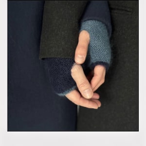 Peut inclure: Gros plan sur deux mains jointes, portant des mitaines bleues. Les gants sont un mélange de tissu tricoté bleu clair et bleu foncé. L'arrière-plan présente des vêtements bleu foncé et noirs.