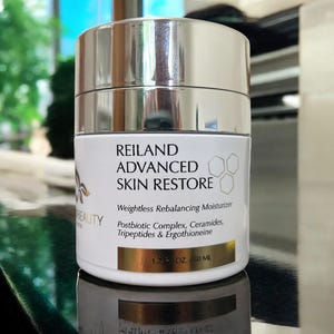 May include: A white and silver jar of Reiland Advanced Skin Restore moisturizer. The label includes the text "Weightless Rebalancing Moisturizer" and "1.7 OZ / 50 ML". The jar is reflected on a dark surface.
