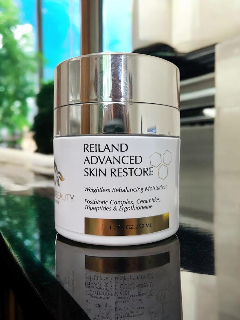 May include: A white and silver jar of Reiland Advanced Skin Restore moisturizer. The label includes the text "Weightless Rebalancing Moisturizer" and "1.7 OZ / 50 ML". The jar is reflected on a dark surface.