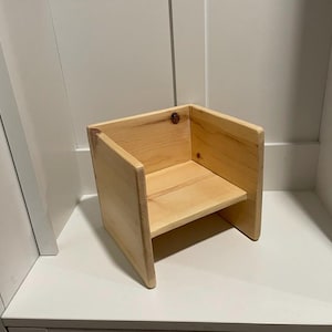 May include: A small, unfinished wooden chair with a simple design. The chair has a seat and back, and is made of light-colored wood. The chair is likely handmade and suitable for a child or as a decorative item.