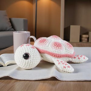 May include: A crocheted turtle toy in white with pink detailing. The turtle features a white shell with pink spots, a pink border, and pink detailing on its flippers. The turtle's head is white with a large, dark eye.