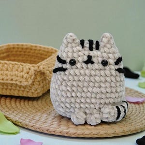 May include: A handmade, plush cat toy in a light gray color with black stripes and facial details. The cat is sitting on a woven mat, with a crocheted basket in the background. The cat toy is a cute, soft, and cuddly item.