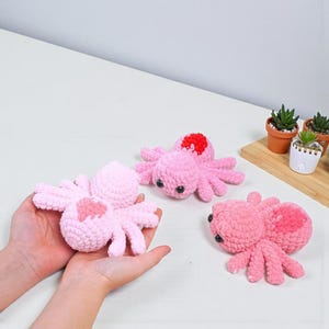 Op de afbeelding: Drie handgemaakte roze gehaakte spinnen met zwarte ogen en rode of roze accenten. Eén spin wordt in open handen gehouden. De spinnen staan op een wit oppervlak, met kleine potplanten op de achtergrond.