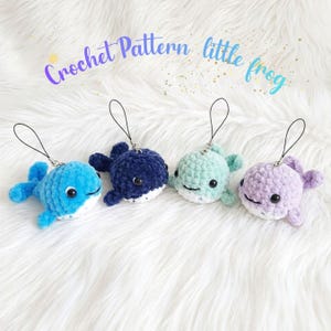 May include: Four small, plush, crocheted whale charms in blue, navy, mint green, and lavender. Each whale features black safety eyes, a white underbelly, and a black cord loop for hanging. The text Crochet Pattern little frog is visible.