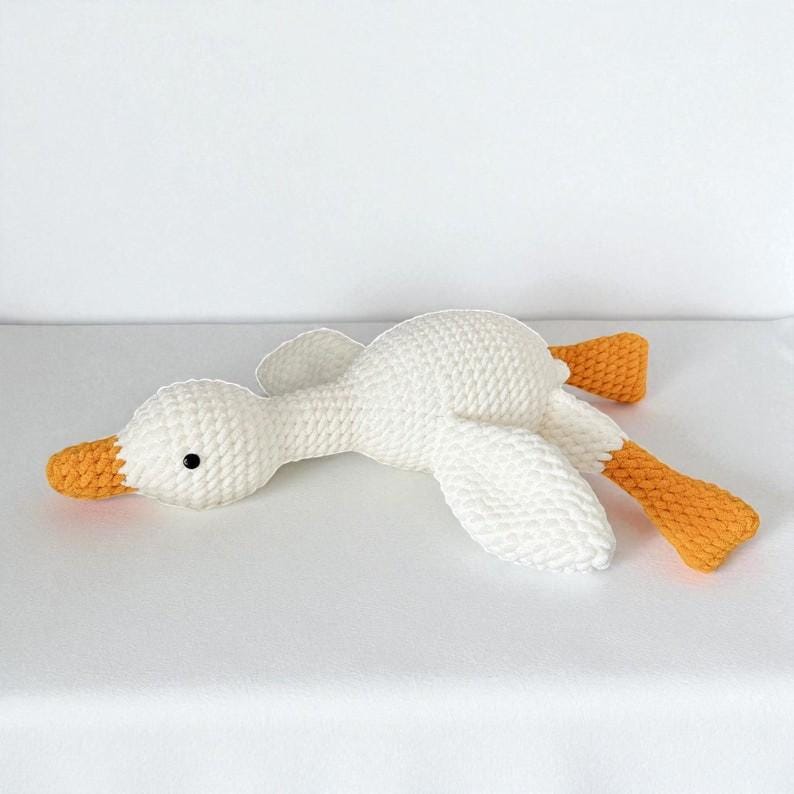 May include: A white crocheted stuffed goose toy with a textured yarn surface. The goose features a bright orange beak and orange webbed feet, with a small black bead used for the eye. The toy is positioned in a horizontal, resting pose.