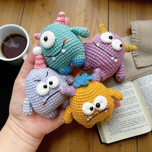 Könnte beinhalten: Vier farbenfrohe, gehäkelte Monstertiere in Blau, Lila, Gelb und Grün. Jedes hat ein einzelnes Auge, einen kleinen Mund und ein Horn oder einen Hut. Die Spielzeuge werden in einer Hand gehalten.