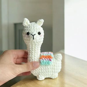 Op de afbeelding: Een handgemaakte witte gehaakte lama met een kleurrijk dekendesign. De lama heeft zwarte ogen, een zwarte neus en kleine oren. De lama is een schattig, zacht speelgoed.