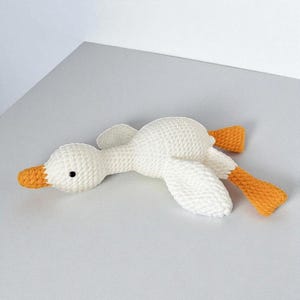 May include: A handmade crochet stuffed animal in the shape of a white goose. The goose features a textured white yarn body, a bright orange beak, and orange webbed feet. It has a single small black bead eye visible on the side of its head.