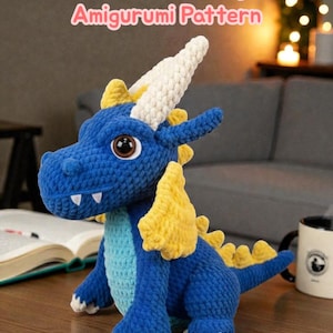 Könnte beinhalten: Ein blauer Häkeldrachen-Amigurumi mit gelben Akzenten, weißen Hörnern und großen braunen Augen. Der Text "Crochet Fire Dragon Amigurumi Pattern" wird über dem Drachen angezeigt. Der Drache sitzt auf einer Holzoberfläche.