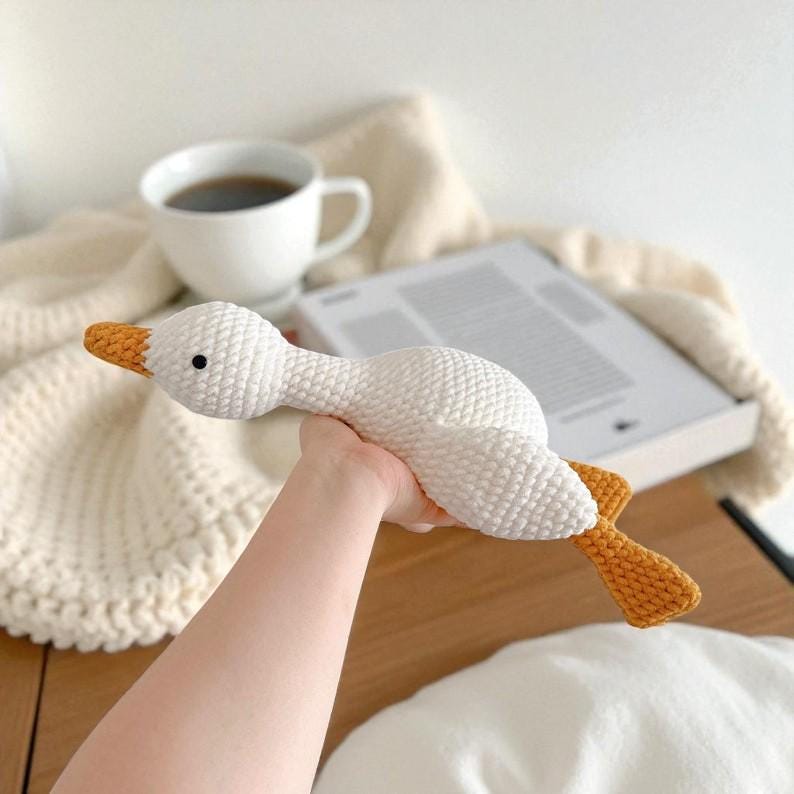 May include: A white crocheted goose toy with a textured yarn finish, featuring a bright orange beak and orange webbed feet. The goose has a small black bead eye and a long neck, crafted with a visible stitch pattern throughout the body.