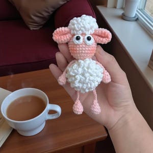 May include: A crocheted sheep toy, featuring white wool and pink details. The sheep has large eyes and small pink legs. It is held in a hand, with a cup of tea on a wooden table in the background. A charming, handmade item, perfect as a gift or decoration.