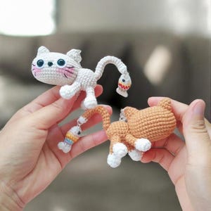May include: Two crochet cat figurines. One is white with pink cheeks and a fish dangling from its tail. The other is orange with white paws. Both are handmade and have a whimsical design.