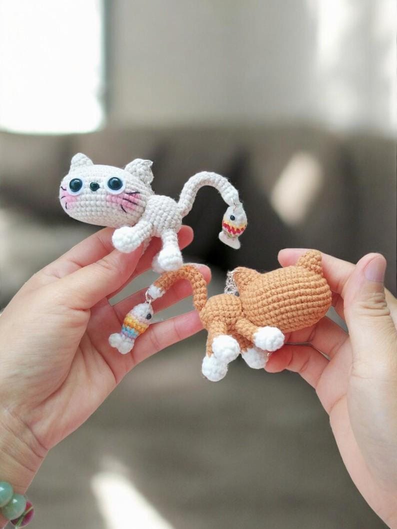 May include: Two crochet cat figurines. One is white with pink cheeks and a fish dangling from its tail. The other is orange with white paws. Both are handmade and have a whimsical design.
