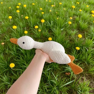May include: A white crocheted goose toy with a textured yarn surface, featuring a bright orange beak and orange webbed feet. The goose has a small black bead eye and is crafted with a soft, plush-like stitch pattern throughout its body.