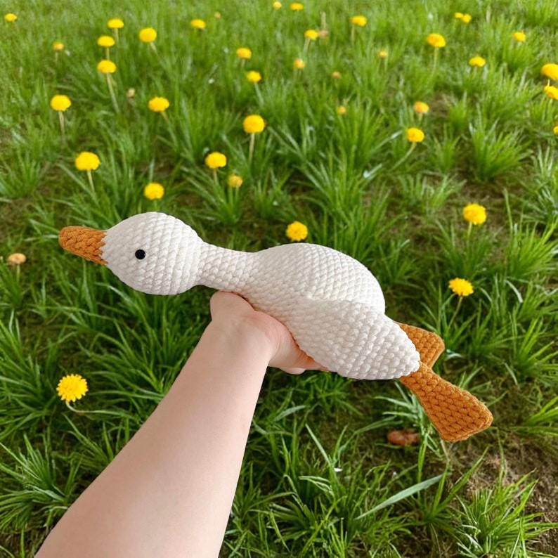 May include: A white crocheted goose toy with a textured yarn surface, featuring a bright orange beak and orange webbed feet. The goose has a small black bead eye and is crafted with a soft, plush-like stitch pattern throughout its body.