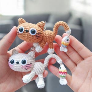 Puede incluir: Dos llaveros de gato de ganchillo. Uno es marrón con rayas negras y patas blancas, el otro es blanco con mejillas rosas. Cada gato tiene ojos negros grandes y una cola con un pequeño pez colorido adjunto.